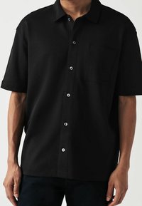 Musta lyhythihainen nappikaulusshirt, joka on valmistettu tekstuurikankaasta. Siinä on yksi rintatasku ja hopeanväriset napit edessä.