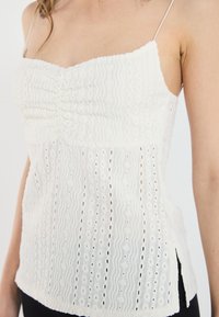 Fehér camisole top állítható spagetti pántokkal, texturált anyag, hullámos mintákkal és gyöngyös részletekkel, oldalsó hasítékokkal a kényelmesebb viselés érdekében.