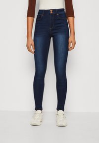 Džínové skinny kalhoty vysokého pasu tmavě modré barvy, vyrobené z denimu, se dvěma předními kapsami a kontrastním oranžovým šitím, nošené s bílými teniskami.