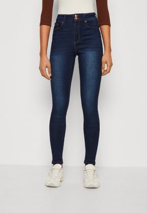 High-waisted dark blue skinny jeans made of denim, featuring two front pockets and contrasting orange stitching, worn with white sneakers.