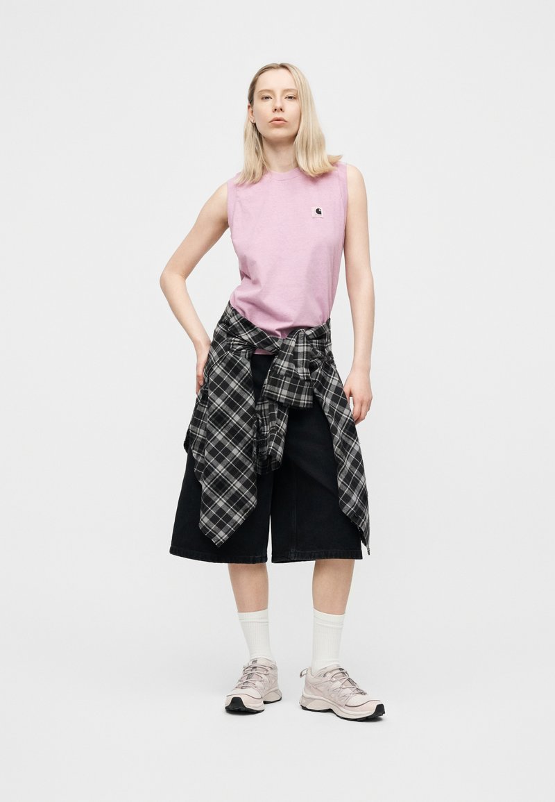 Woman standing with hand on hip wearing sleeveless pink top, black knee-length shorts, plaid shirt tied around waist, white socks, and sneakers.