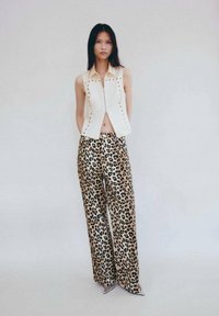 Top bianco senza maniche con zip frontale e applicazioni di borchie, abbinato a pantaloni larghi con stampa leopardata. La modella è in piedi davanti a uno sfondo chiaro.
