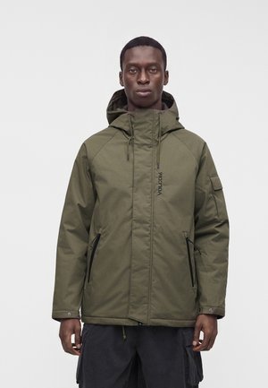 Homme portant une veste à capuche vert olive avec fermeture éclair devant et un pantalon cargo noir sur fond blanc uni