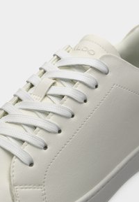 Gros plan d'une basket en cuir blanc avec des lacets blancs et des détails de couture sur un fond clair.