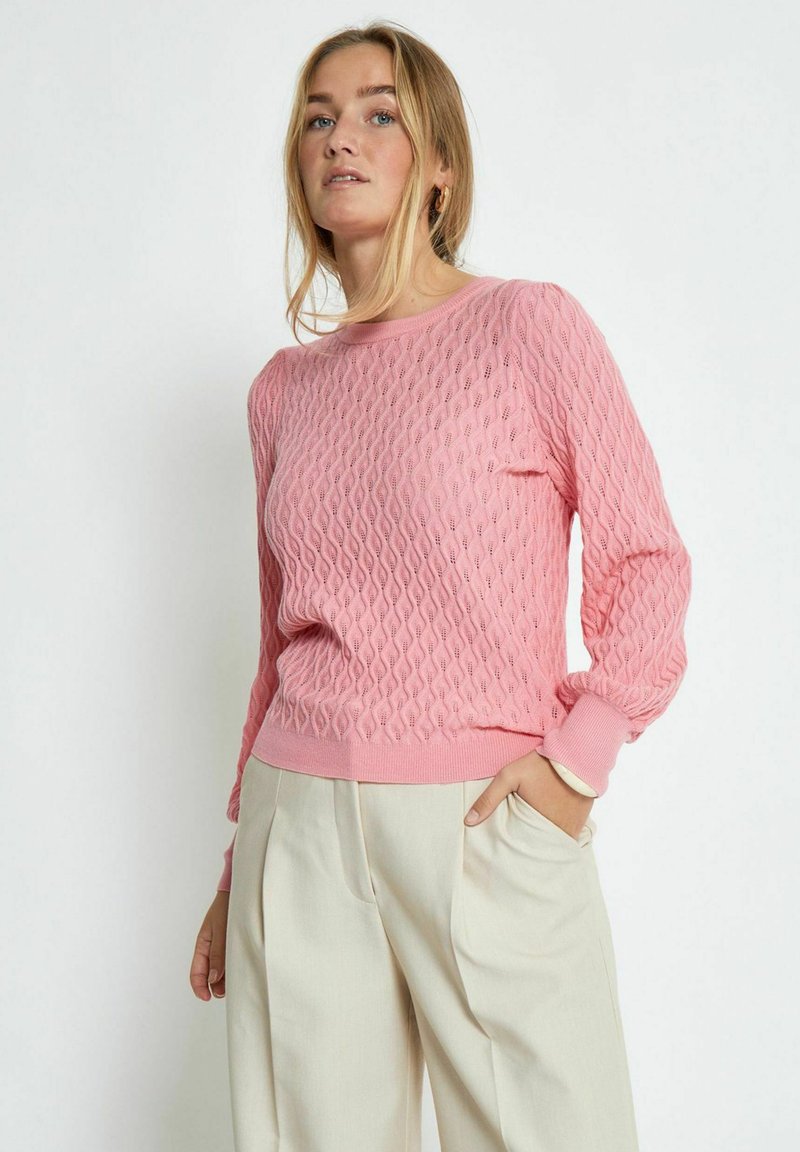 Maglione rosa con trama a diamante, polsini e orlo a costine. Indossato con pantaloni color crema chiaro, che mostrano un fitting rilassato.