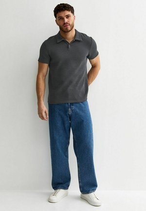 Mężczyzna stojący na tle białej ściany, ubrany w ciemnoszary polo z krótkim rękawem, luźne niebieskie dżinsy i białe trampki, trzymający jedną rękę za plecami.