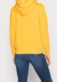 Gelbes Hoodie mit Kordelzugkapuze, gerippten Bündchen und Saum, kombiniert mit blauen Jeans aus Denim, die ein kleines Markenlogo auf der Gesäßtasche tragen.