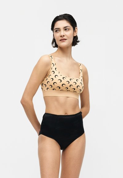 Γυναίκα με κοντά σκούρα μαλλιά που φοράει ροδακινί αθλητικό σουτιέν με μαύρα ημισεληνοειδή σχέδια και ψηλόμεσο μαύρο κάτω μέρος, στέκεται μπροστά σε λευκό φόντο.