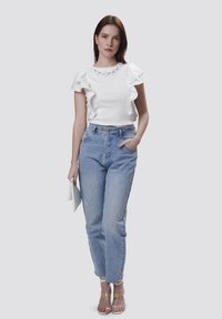 Hvid kortærmet top med bølgede kanter, parret med højtaljede lyseblå jeans. Gennemsigtige hælede sandaler og holder en lyseblå clutch.