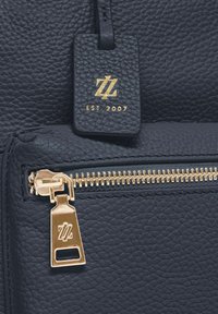 Nahaufnahme einer marineblauen strukturierten Lederhandtasche mit goldfarbenem Reißverschluss und passendem Lederanhänger, der mit einem goldenen Logo und "EST. 2007." geprägt ist.