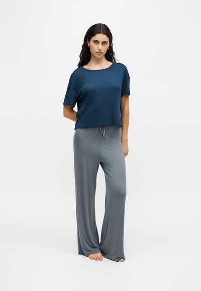 Jeune femme aux longs cheveux foncés portant un haut ample à manches courtes bleu marine et un pantalon large gris de détente, debout pieds nus sur un fond uni.
