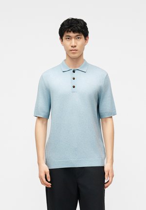 Jonge man die een lichtblauw, gebreid poloshirt met korte mouwen en drie bruine knopen draagt, staand tegen een effen witte achtergrond.