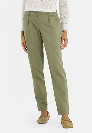 Mujer con pantalones verde oliva de pierna recta, blusa blanca de manga larga con bordados calados y zapatos planos beige con detalles calados.
