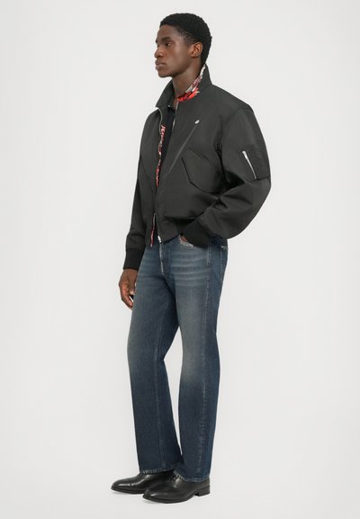 Black bomber jacket with a high collar, zip front, and two large front pockets. Worn with blue jeans and black shoes. Patterned inner lining visible.