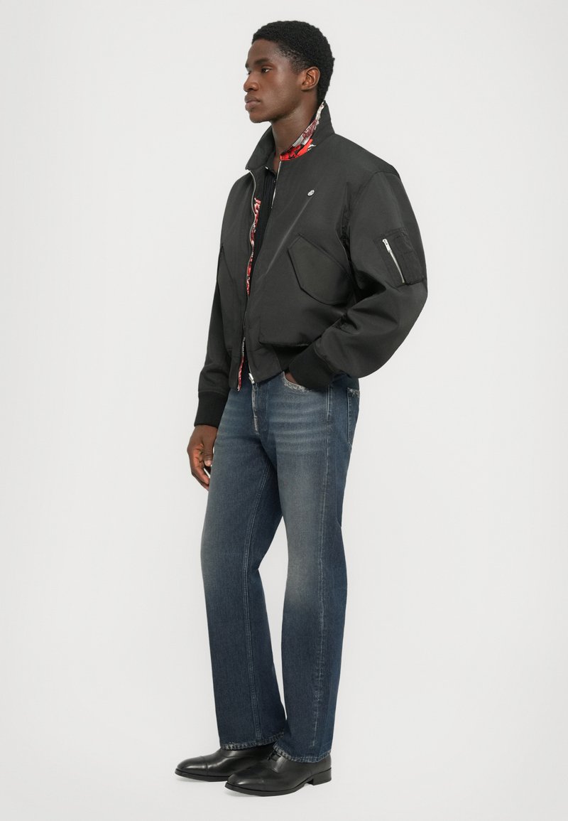 Giubbotto bomber nero con collo alto, zip frontale e due grandi tasche anteriori. Indossato con jeans blu e scarpe nere. Fodera interna a fantasia visibile.