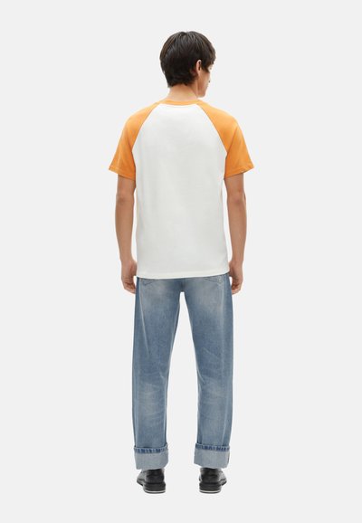 Kurzärmliges Raglan-T-Shirt mit orangefarbenen Ärmeln, weißem Körper und lässiger Passform, kombiniert mit hellblauen, hochgekrempelten Jeans und schwarzen Schuhen.