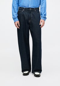 Person mit dunkelblauen, weiten Jeans, weißen und schwarzen Schuhen und einem blauen Langarmshirt mit vertikalen weißen Streifen.