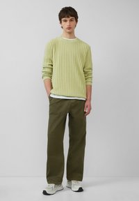 Lichtgroene ribgebreide trui, gelaagd over een wit shirt, gecombineerd met olijfgroene losse broek en beige hardloopschoenen.