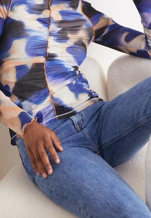 Personne assise sur une chaise blanche portant un haut à manches longues bleu et beige à motif abstrait avec des coutures orange et un jean en denim bleu.