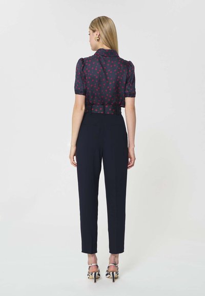 Femme aux cheveux blonds raides portant un pantalon bleu marine, un chemisier bleu marine à manches courtes avec des motifs rouges, et des chaussures à talons hauts avec des brides, debout de dos.