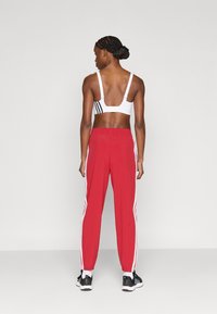 Λευκό αθλητικό σουτιέν με μαύρες ρίγες στην πλάτη, συνδυασμένο με κόκκινο αθλητικό παντελόνι με λευκές πλευρικές ρίγες και μαύρα αθλητικά παπούτσια.