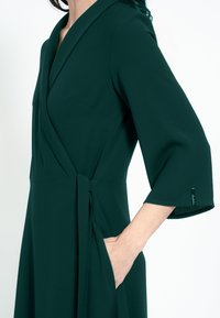 Zelené zavinovacie šaty s hlbokým výstrihom do V, širokými rukávmi a opaskom. Majú bočnú kapsu a hladký povrch. Na rukáve je štítok.