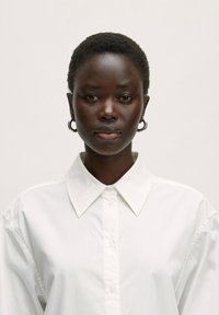 Weißes Hemd mit Knopfleiste, aus glattem Stoff. Das Model trägt runde silberne Ohrringe und hat eine kurze, natürliche Frisur.