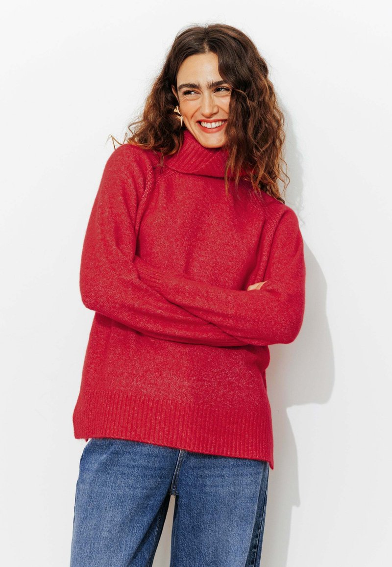 Femme souriante avec des cheveux bouclés bruns, portant un pull col roulé rouge vif et un jean bleu, se tenant les bras croisés devant un fond blanc.