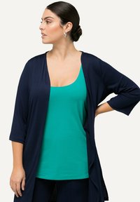 Cárdigan de canalé en color azul marino con mangas tres cuartos sobre una camiseta de tirantes en un tono sólido de teal. El cárdigan presenta un diseño de apertura frontal.