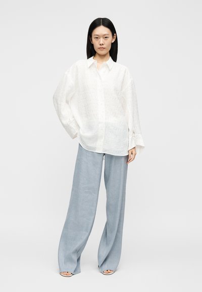 Femeie stând cu mâna în buzunar, purtând o cămașă largă albă brodată cu nasturi și pantaloni largi bleu deschis cu sandale cu degetele goale.