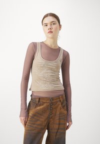 Débardeur beige avec motif d'écriture, superposé sur une chemise à manches longues mauve. Pantalon en velours côtelé brun avec rayures verticales et fermeture à cinq boutons.