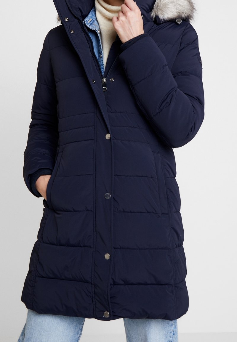 Piumino blu navy con cappuccio in pelliccia staccabile, fantasia trapuntata, chiusure a scatto frontali e tasche laterali. Indossato sopra un maglione beige.