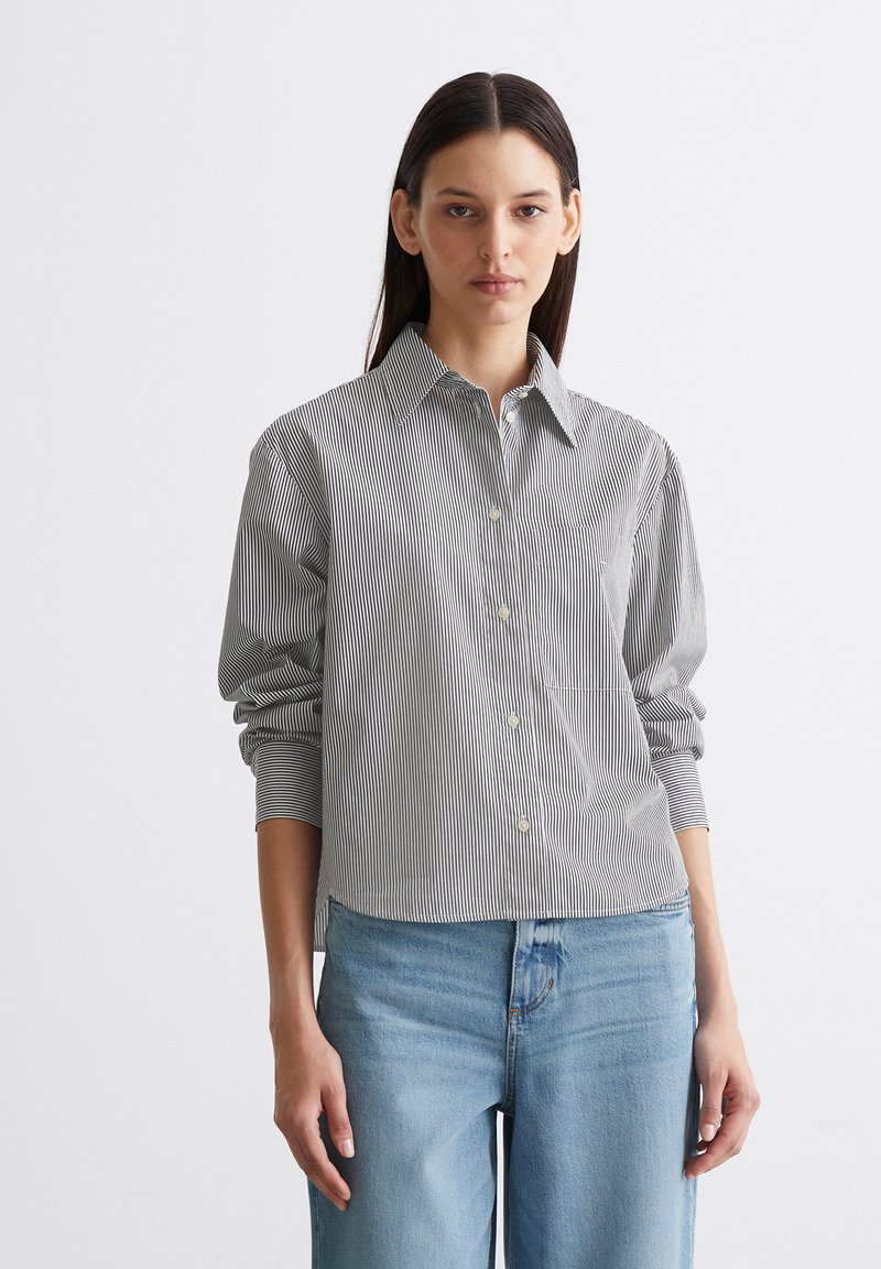 Gestreifte Hemdbluse in Schwarz und Weiß mit klassischem Kragen, langen Ärmeln und lässiger Passform. Kombiniert mit hellblauen Jeans.