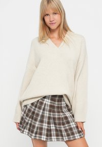 Maglione beige oversize con scollo a V, abbinato a una mini gonna plissettata a quadri nei toni marrone e bianco. Tessuto morbido e testurizzato.