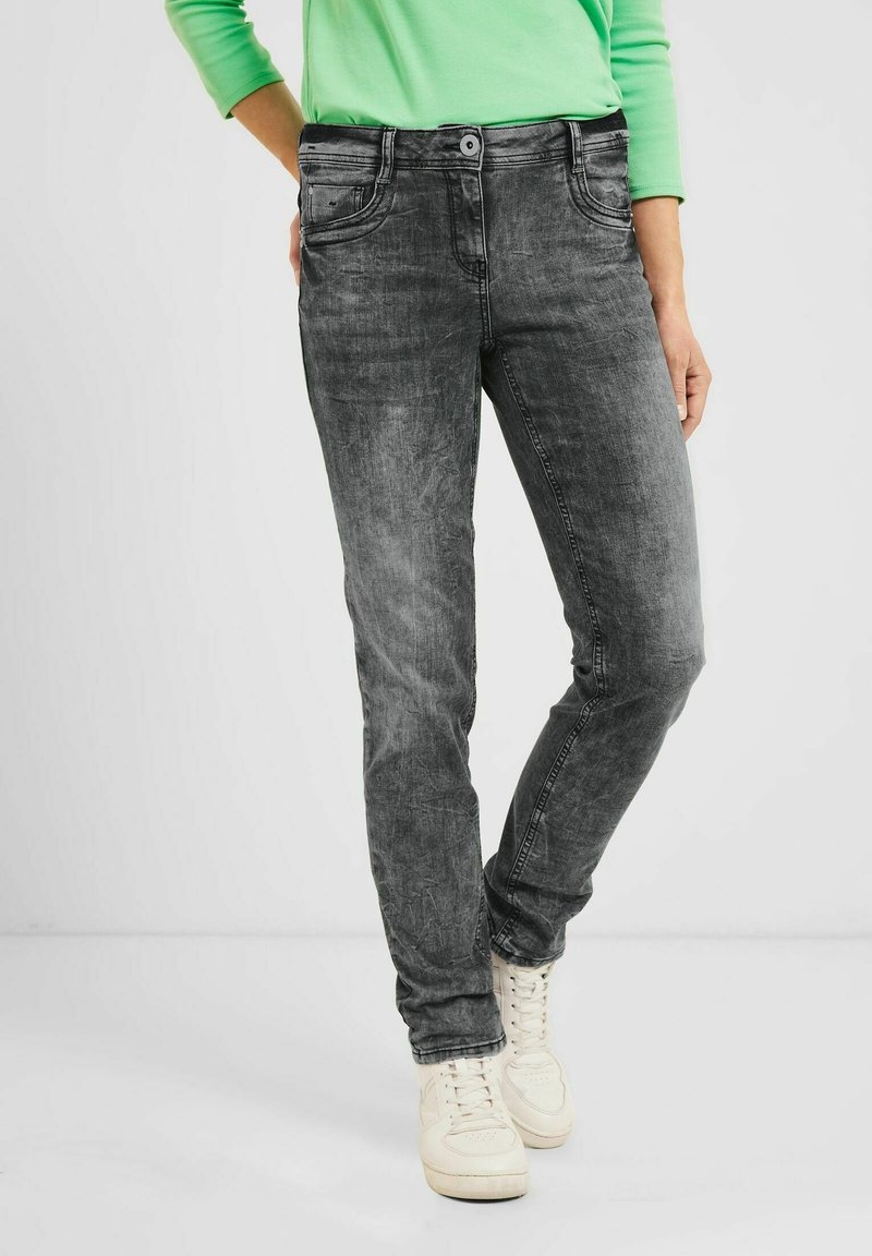 Jeans de denim gris con un acabado ligeramente desgastado. Presenta un diseño clásico de cinco bolsillos, pasadores para cinturón y un cierre de botón. Combinados con zapatillas blancas.