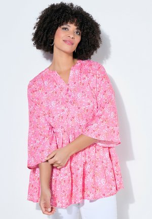 Kobieta o kręconych włosach, nosząca kolczyki-koła i luźną różową bluzkę w kwiaty z rękawami 3/4, pozuje na białym tle.