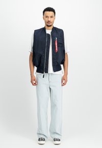 Chaleco de bombardero navy con cremallera, bolsillo frontal y detalle de etiqueta roja. Llevado sobre una camiseta blanca. Combinado con jeans de pierna ancha de color azul claro y zapatos negros.