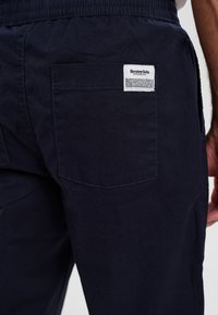 Mørkeblå bukser med elastisk talje, sidelommer og en baglomme med en hvid etiket, der viser mærkeoplysninger.