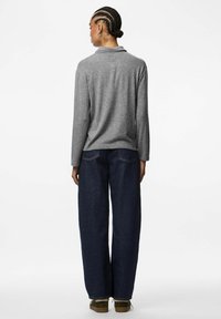 Osoba s copy v vlasů, oblečená v šedém svetru s dlouhým rukávem a tmavých širokých džínách, stojící zády k objektivu na jednobarevném pozadí.