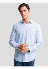 Mann trägt ein hellblaues Hemd mit Knöpfen und Brusttasche sowie dunkle Jeans, steht vor einem einfarbigen grauen Hintergrund und hält sich eines der Handgelenke.