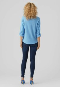 Blusa azzurro chiaro, con texture e maniche arrotolate, abbinata a jeans skinny blu scuro e sandali con tacco blu. Vista posteriore.