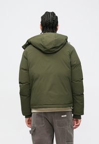 Casaco acolchoado verde-oliva com capuz, apresentando uma textura suave e punhos elásticos, usado sobre calças cargo cinzentas.