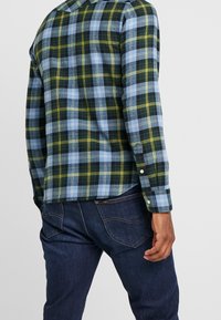 Grön, blå och gul rutig flanellskjorta med knäppta ärmslut, bärs över mörkblå jeans. Mjuk textur, rak skärning och en krage.