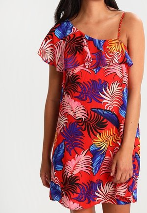 Femme portant une robe rouge à une épaule avec des motifs de feuilles tropicales bleues, roses, violettes, jaunes et noires.