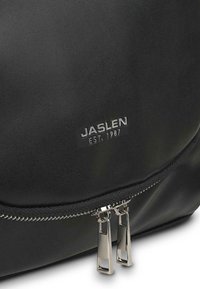 Mochila de cuero negro con cierre con cremallera, herrajes en tono plateado, logo grabado "JASLEN EST. 1987" y textura suave.