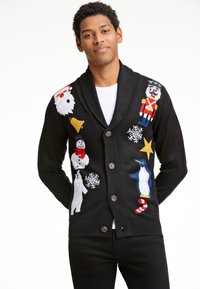 Giovane uomo che indossa un cardigan nero con bottoni e fantasie natalizie tra cui Babbo Natale, pupazzo di neve, soldatino, pinguino, orso polare e fiocchi di neve.