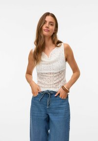 Haut sans manches blanc avec un design en dentelle florale texturée, présentant un décolleté drapé, associé à un jean en denim bleu ample.