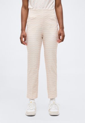 Άτομο που φοράει ανοιχτό μπεζ ψηλόμεσα παντελόνια με σχέδια και μπροστινές τσέπες, καθώς και λευκά αθλητικά παπούτσια, στέκεται μπροστά σε ένα απλό ανοιχτό φόντο.