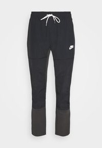 Zwarte Nike joggers met een witte trekkoord tailleband en een wit Nike-logo op de bovenlinke dij, taps toelopende benen met verschillende stoffenpanelen onder de knieën.