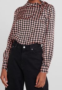 Långärmad blus i rosa och svart houndstooth-mönster, med en slipsdetalj vid halsringningen och mjukt tyg; kombinerat med svarta högmidjade jeans.